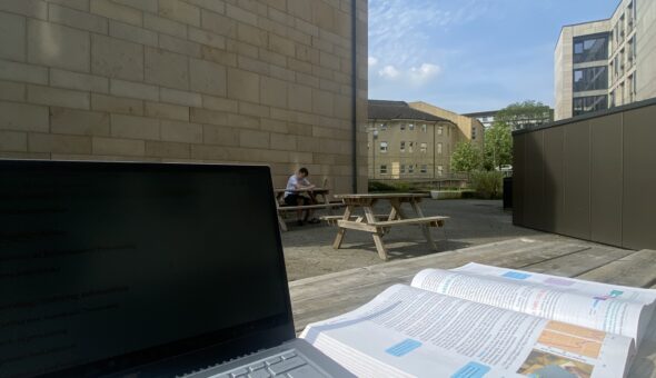 Image of a desk outside. On the desk, in the foreground is a book and laptop. In the background is a sunny building courtyard