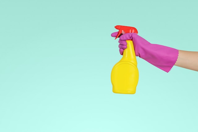 An extended arm in a pink rubber glove holds a yellow cleaning spray bottle.