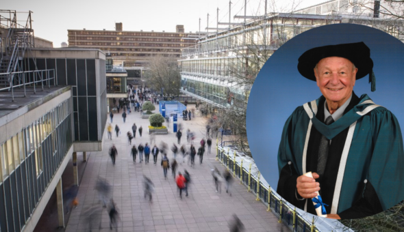 An old man in a ceremonial graduation cap and gown holding a degree and smiling against of backdrop of the University of Bath Parade street.