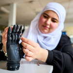 A young female student wearing a hijab adjusts a robotic hand in a lab.