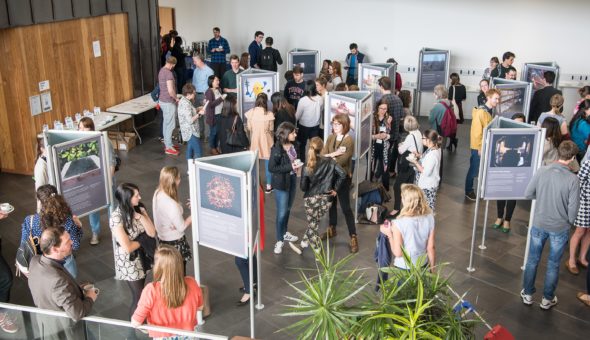 An exhibition Hall with a crowd of people of different genders and ethnicities standing around the exhibition stands viewing the exhibition and in discussion with other visitors