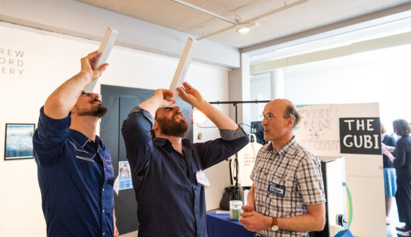 Two men looking up through small paper tubes while another man talks to them.