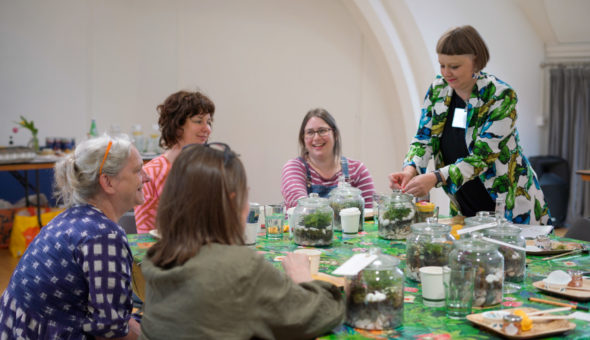 Five people sit around a table creating terrariums together. They appear deep in conversation.